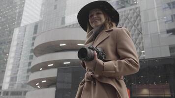 aantrekkelijk jong blond vrouw met een camera in haar handen in de stad straat. actie. elegant vrouw in bruin jas en zwart hoed glimlachen en poseren Aan een winter dag Aan de achtergrond van modern gebouwen. video