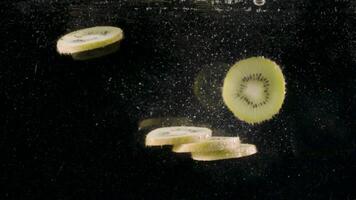 Slices of kiwi fruit drop into water full of bubbles in slow motion. Action. Close up of cut tropical fruit falling underwater isolated on black background. video