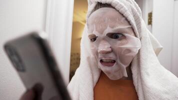 Portrait of a young girl with a towel on her head and a fabric face mask on her head looking at smartphone. Action. Teenager girl looking shocked and angry, getting bad news. video