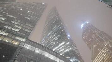 Modern skyscrapers in snowfall. Action. Business center with glowing skyscrapers during snowfall at night. Beautiful business high-rises on background of cloudy sky with snow at night video