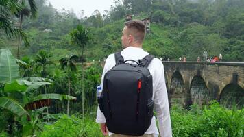 Man in jungle near bridge. Action. Man with backpack looks back at hiking trail in jungle. Handsome man on top of ancient stone bridge in jungle video