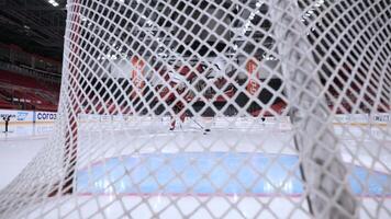 Hockey player trains alone at rink. Action. View through netting of gate at rink. Hockey player trains alone to score puck at rink. Slow motion video