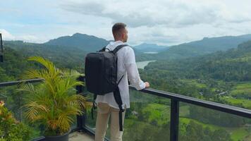 Man with view of tropical mountain valley. Action. Man on observation deck in tropical mountains. Man with backpack on sightseeing attraction overlooking green mountains video