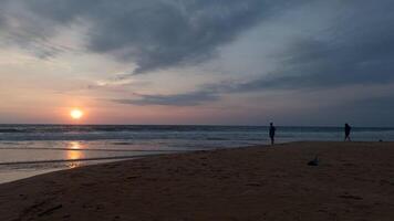 personas son en pie en arena con ver de mar puesta de sol. acción. personas Mira a puesta de sol desde arenoso costa. hermosa paisaje con personas en arena en antecedentes de olas y ajuste Dom video
