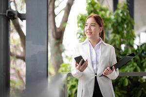 asiático negocio mujer hermosa con inteligente teléfono y documento archivo, asiático mujer con teléfono inteligente caminando Vamos a trabajar. preparación para el trabajando día foto