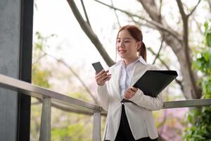 asiático negocio mujer hermosa con inteligente teléfono y documento archivo, asiático mujer con teléfono inteligente caminando Vamos a trabajar. preparación para el trabajando día foto