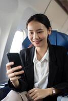 Asian young businesswoman successful or female entrepreneur in formal suit in a plane sit in a business class seat and uses a smartphone during flight. Traveling and Business concept photo