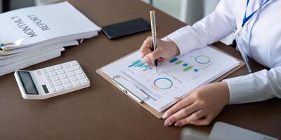 Finance concept. Close up businesswoman hold a graph pen and writing report, and memo, and analyzing business documents with a laptop computer photo