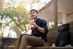 Asian businessman with reusable eco friendly ecological cup using laptop and sitting outside the office building. Eco friendly, sustainable lifestyle concept photo