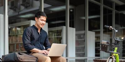 Asian businessman with bicycle using laptop and sitting outside the office building. man commuting on bike go to work. Eco friendly vehicle, sustainable lifestyle concept photo