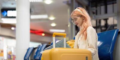 contento joven mujer asiático es sentado en aeropuerto cerca maleta y leyendo mapa foto