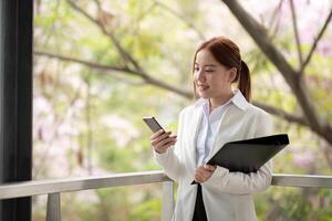 asiático negocio mujer hermosa con inteligente teléfono y documento archivo, asiático mujer con teléfono inteligente caminando Vamos a trabajar. preparación para el trabajando día foto