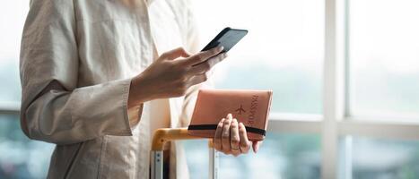 turista mujer utilizando móvil teléfono inteligente y participación pasaporte con maleta de viaje Entre murga para vuelo en aeropuerto Terminal, vuelo cheque en, turista viaje viaje concepto foto