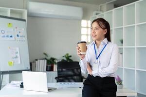 Young successful employee business woman hold craft paper cup coffee stand at desk with laptop pc computer at office. Achievement career concept photo