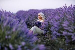 Blonde woman poses in lavender field at sunset. Happy woman in white dress holds lavender bouquet. Aromatherapy concept, lavender oil, photo session in lavender