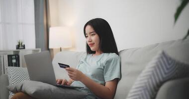 selectivo enfocar, joven asiático mujer sentado en sofá participación crédito tarjeta haciendo en línea pago para compra en web Tienda utilizando ordenador portátil. foto