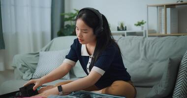 Selective focus of Asian teenager woman wear wireless headphones sitting on sofa packing travel luggage with personal items for traveling trip, Preparation travel suitcase at home. photo