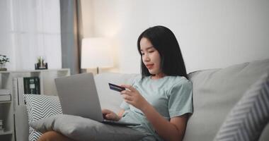 selectivo enfocar, joven asiático mujer sentado en sofá participación crédito tarjeta haciendo en línea pago para compra en web Tienda utilizando ordenador portátil. foto
