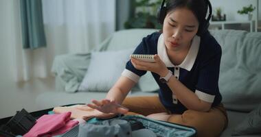 Selective focus of Asian teenager woman wear wireless headphones sitting on sofa making checklist of things to pack for travel, Preparation travel suitcase at home. photo