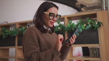een vrouw in bril is leunend Aan een bureau en gebruik makend van haar telefoon video