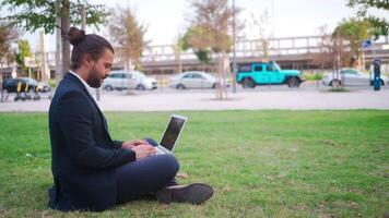 ein Mann im ein passen Sitzung auf das Gras mit ein Laptop video