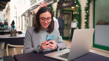 uma mulher dentro óculos sentado às uma mesa com uma computador portátil e célula telefone video