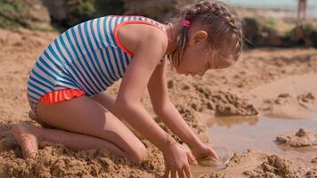 un' poco ragazza nel un' a strisce costume da bagno è giocando con sabbia nel il spiaggia video