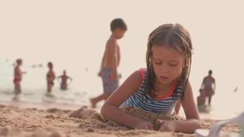 un' poco ragazza nel un' a strisce costume da bagno è giocando con sabbia nel il spiaggia video