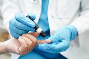 Asian doctor using lancet pen on senior patient finger for check sample blood sugar level to treatment diabetes. photo