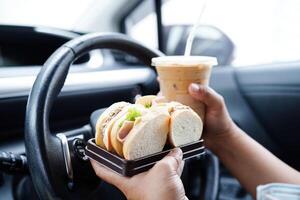 Asian woman driver hold and eat hamburger in car, dangerous and risk an accident. photo