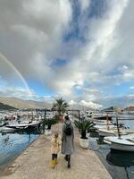 Budva, Montenegro - 25 december 2022. Mom with a little girl walk along the pier against the backdrop of a rainbow over the mountains photo