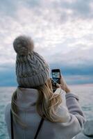 niña soportes en el apuntalar y dispara el puesta de sol terminado el mar con un teléfono inteligente espalda ver foto
