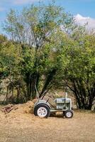 Small tractor stands near the hay in a pasture near green trees photo