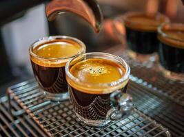 cerca arriba de Café exprés Disparo con café crema y profesional café máquina mientras preparando dos espressos Disparo vaso en un café tienda. foto