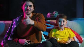 zwei Brüder sind Sitzung zusammen, eingewickelt im ein amerikanisch Flagge, Aufpassen Fernseher und Jubel auf ihr Liebling Sport Team. Sie sind Lachen und genießen jeder Andere Unternehmen. Mitte Schuss. 4k video