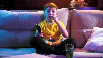 een jong jongen horloges televisie in de avond, zittend Aan de bank, aan het eten chips van een schaal. midden schot. 4k video