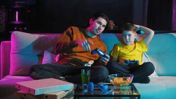 Older brother helps the younger one by explaining how to play a game on the console using the wireless joystick at night in the living room, while snacks, soda, and pizza are placed on the table before them. Mid shot. 4k video