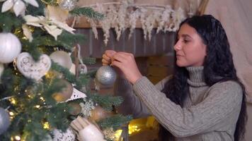 familie kerstmis, pret partij, blijven Bij huis, nieuw jaar viering. jong vrouw siert Kerstmis boom met mooi glimmend bal en glimlacht video