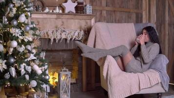 família Natal, Diversão festa, fique às lar, Novo ano celebração. uma mulher bebidas chá a partir de uma caneca enquanto sentado em a poltrona perto uma Natal árvore video