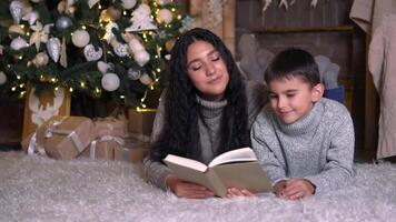 famiglia Natale, divertimento festa, restare a casa, nuovo anno celebrazione. mamma è lettura un' libro per sua figlio mentre dire bugie su il pavimento vicino il Natale albero video