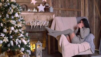 familj jul, roligt fest, stanna kvar på Hem, ny år firande. en kvinna i en stickat Tröja sitter i en stol nära en jul träd och drycker te och muggar video