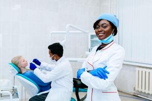 joven sonriente africano americano hembra dentista en frente de dentista comprobación dientes a paciente a clínica. dental clínica concepto. foto