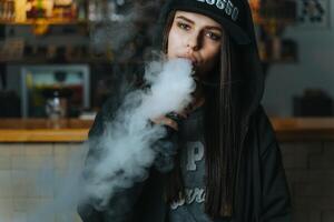 Young pretty woman in cap smoke an electronic cigarette at the vape shop. Hip-hop style. Closeup. photo
