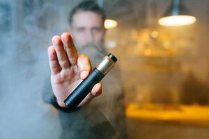 Young beard man show vaping device on his outstretched hand through a cloud of steam. Selective focus. Vaping concept. Copy space. photo