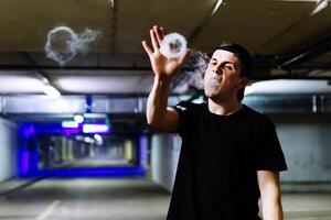 Man in cap smoke an electronic cigarette and releases clouds of vapor performing various kind of vaping tricks photo