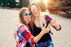 retrato de un contento dos sonriente muchachas haciendo selfie foto en teléfono inteligente urbano antecedentes. el noche puesta de sol terminado el ciudad.