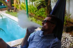 hombre descansando en un hamaca y vaping y dejando apagado vapor desde un electrónico cigarrillo foto