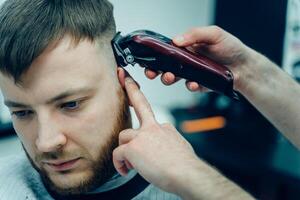 Male haircut with electric razor. Barber makes haircut for client at the barber shop by using hairclipper. Man hairdressing with electric shaver. photo