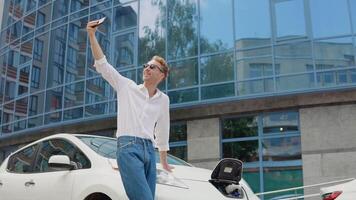 eleganta modern ung lockigt man står nära ett elektrisk bil laddning och tar en selfie med hans smartphone video