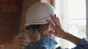 A female architect stands with her back to the camera and looks out the window at the construction site. Woman builder adjusts protective helmet on her head video